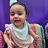 baby, child, girl, smiling, pink_clothing, white_bib, bow, curly_hair, colorful_chair, purple_wall, indoor, cute, happy, toddler, portrait, playful, small_earrings, skin, face, person