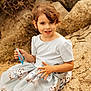 beach_rocks, brown_hair, candid, candy, child, earrings, eyes, girl, hands, kitten_print, nature, outdoors, portrait, rocks, short_sleeves, sitting, skirt, smile, sunlight, warm_tones
