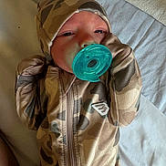 Sutton is registered to the contest to win money with this photo: baby, pacifier, camouflage, clothing, hood, infant, sleepy, bed, blanket, soft, cute, newborn, person, portrait, lying_down, cozy, child, small, warm, comfort