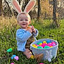 baby, basket, blue_cardigan, bunny_ears, child, cute, easter_eggs, grass, greenery, happy, nature, outdoor, playful, seasonal, sitting, smiling, spring, sunlight, toy, trees