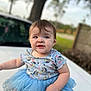 Theresa is registered to the contest to win money with this photo: baby, child, blue_tutu, shirt, outdoor, nature, tree, fence, white_surface, smile, curious, cute, sitting, young_child, person, short_hair, daylight, grass, playful, portrait