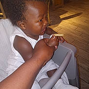 Lan is registered to the contest to win money with this photo: baby, child, high_chair, french_fry, hand, indoor, flooring, curly_hair, white_clothing, sock, feeding, person, table, seat, wood_floor, light, shadow, skin, expression, curiosity