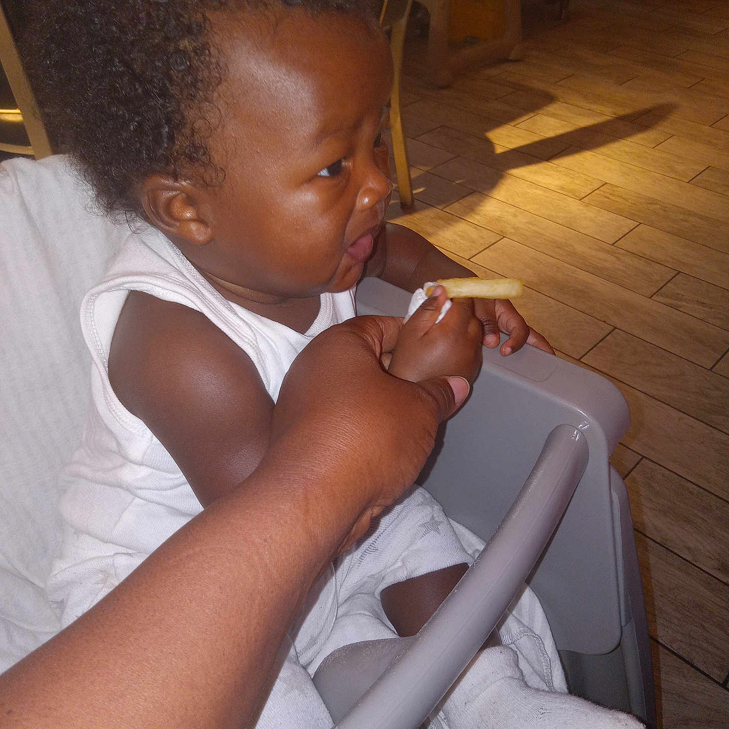 Lan is registered to the contest to win money with this photo: baby, child, curiosity, curly_hair, expression, feeding, flooring, french_fry, hand, high_chair, indoor, light, person, seat, shadow, skin, sock, table, white_clothing, wood_floor