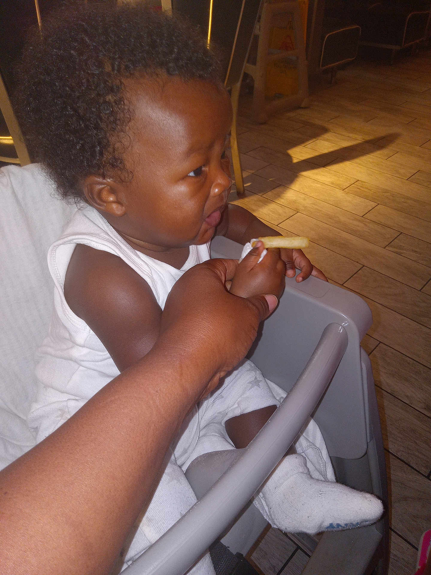 Lan is registered to the contest to win money with this photo: baby, child, high_chair, french_fry, hand, indoor, flooring, curly_hair, white_clothing, sock, feeding, person, table, seat, wood_floor, light, shadow, skin, expression, curiosity