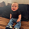 baby, child, smiling, sitting, blue_eyes, black_shirt, jeans, sneakers, bed, pillow, indoor, wooden_wall, cute, happy, portrait, young_child, clothing, footwear, infant, casual