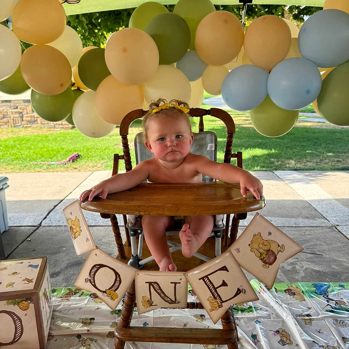 Renezmae is registered to the contest to win money with this photo: baby, high_chair, birthday, balloons, banner, winnie_the_pooh, party, outdoor, child, celebration, wooden_chair, decorations, tablecloth, grass, sunlight, cute, one_year_old, event, childrens_party, infant
