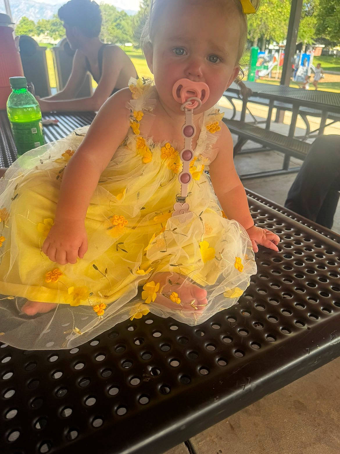 Renezmae is registered to the contest to win money with this photo: baby, bench, casual, child, daytime, girl, grass, green_bottle, nature, outdoor, pacifier, park, person, picnic_table, plastic_bottle, playground, summer, sunlight, trees, yellow_dress