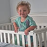 toddler, child, crib, bedroom, white_crib, smiling, blonde_hair, tousled_hair, standing, gripping, railing, indoor, bed, blanket, plush_toy, light_wall, casual_clothing, cute, happy, infant