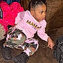 child, girl, couch, pink_sweatshirt, camouflage_pants, black_boots, red_jacket, brown_furniture, indoor, casual_clothing, sitting, curious_expression, hairstyle, earrings, bag, floor, cozy, young, portrait, person