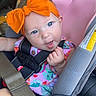 baby, car_seat, orange_bow, blue_eyes, child, seatbelt, colorful_clothing, infant, cheeky_expression, car_interior, safety_harness, cute, portrait, seat, headband, young_child, smiling, person, indoors, closeup