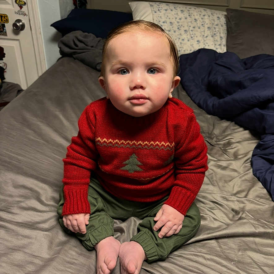 Mylo is registered to the contest to win money with this photo: baby, bed, blanket, casual, child, cozy, cross_legged, cute, face, green_pants, home, indoor, looking_at_camera, pillow, portrait, red_sweater, sitting, sweater, toddler, young_child