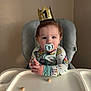 Mylo joined the competition — help win amazing prizes! baby, child, high_chair, pacifier, crown, birthday, snack, face, hands, clothing, onesie, indoor, table, seat, celebration, expression, young_child, cute, portrait, infant