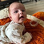 baby, infant, blanket, orange, white_clothing, animal_print, face, eyes, mouth, indoors, floor, wooden_furniture, curious, lying_down, soft_texture, child, cute, person, toddler, cozy