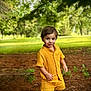 child, toddler, yellow_clothing, shorts, shirt, outdoor, forest, trees, grass, sunlight, nature, greenery, smiling, standing, boy, casual_clothing, daylight, happy, footwear, pine_needles