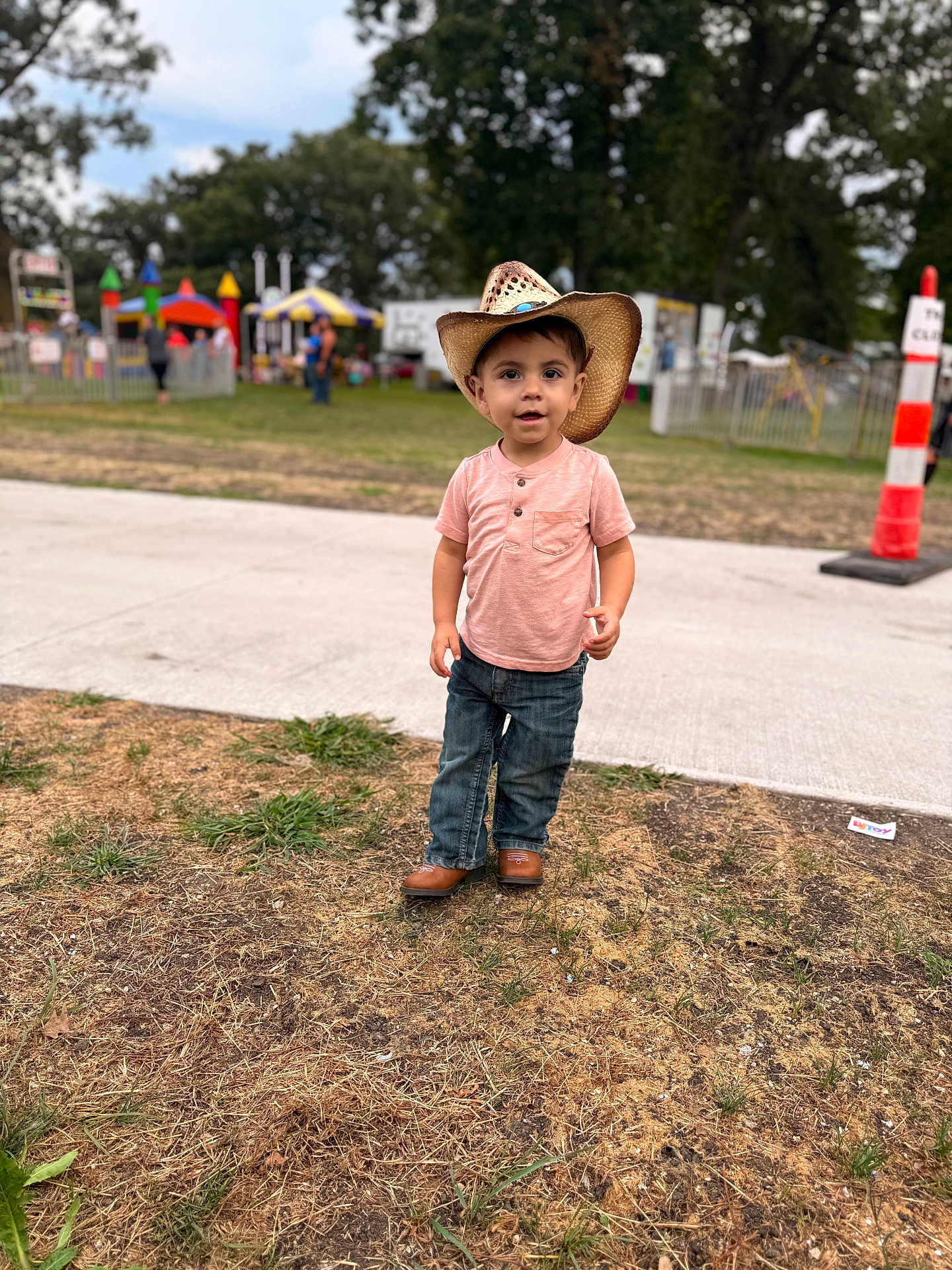 Amelliano is registered to the contest to win money with this photo: toddler, child, cowboy_hat, pink_shirt, jeans, brown_shoes, grass, sidewalk, outdoor, fairground, people, trees, daytime, playground, fence, hat, curious, walking, casual_clothing, event