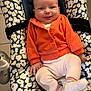 baby, child, infant, smile, orange_jacket, white_pants, baby_seat, patterned_cushion, comfortable, cute, indoors, happy, person, sitting, soft_clothing, small_hands, footwear, playful, portrait, cozy