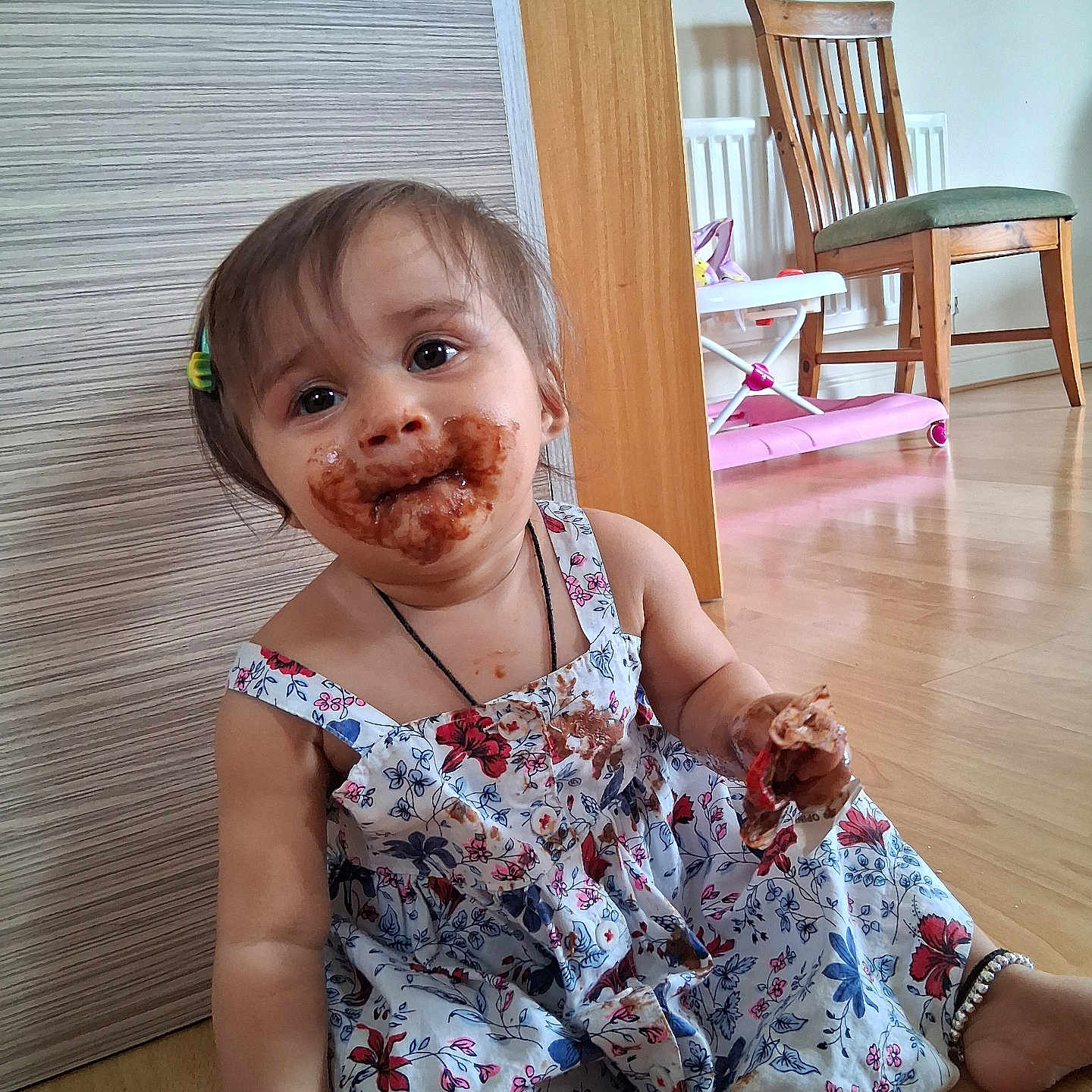 Kriva is registered to the contest to win money with this photo: baby_walker, chair, child, chocolate, face, floor, floral_dress, foot, hair_clip, hand, happy, indoor, messy, necklace, person, sitting, smile, toddler, toy, wooden_floor