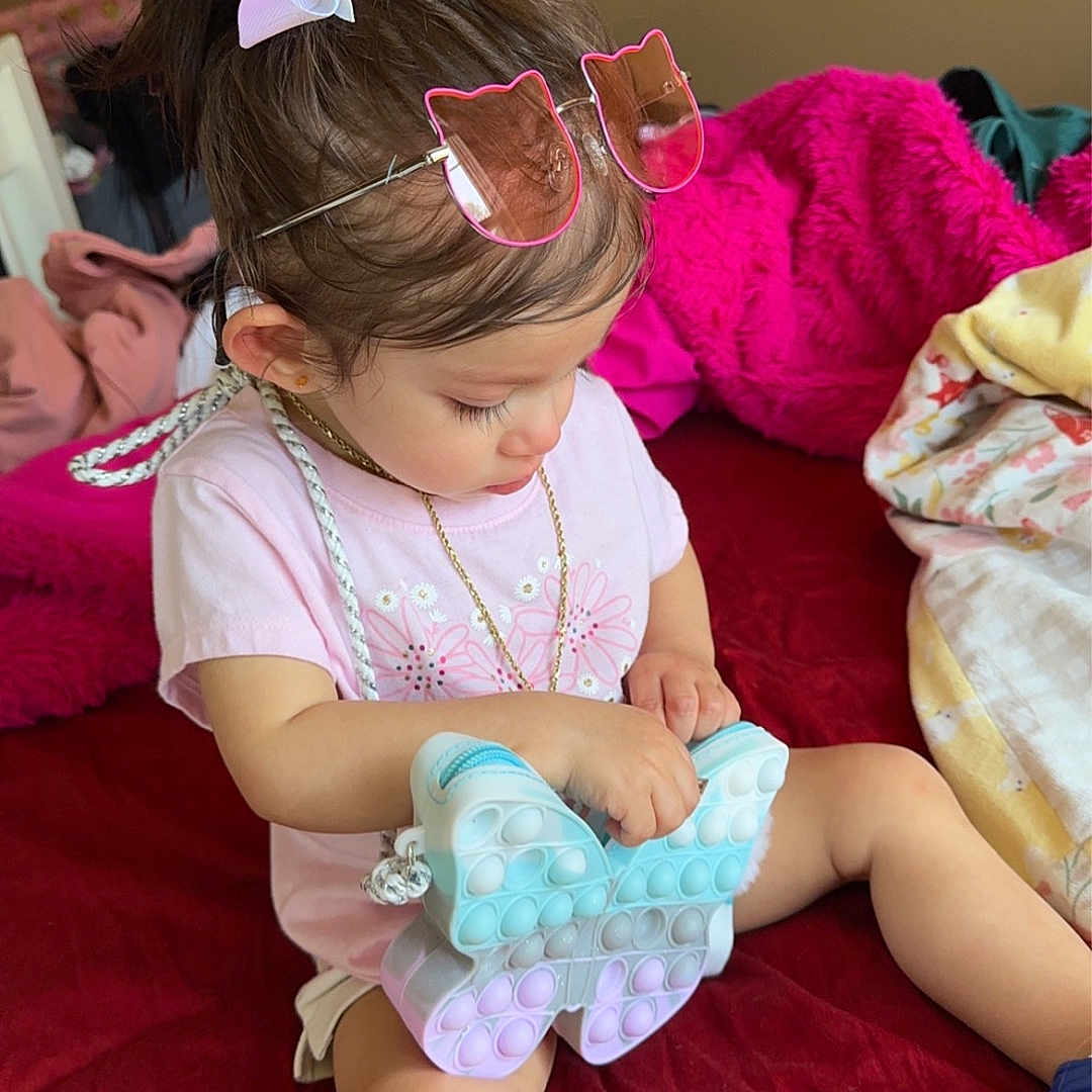 Zairianna Mills joined the competition — help win amazing prizes! toddler, child, toy, pop_it, butterfly, sunglasses, bow, pink_clothing, blue_shoes, bed, blanket, indoors, person, cute, young_child, playing, sitting, accessory, hair_bow, casual