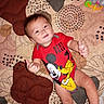 baby, child, infant, smiling, happy, lying_down, thumbs_up, portrait, face, eyes, hair, red_clothing, onesie, cartoon_character, mickey_mouse, pluto, bed, bedspread, toy, indoor