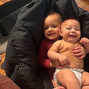 Melody joined the competition — help win amazing prizes! baby, infant, child, smile, diaper, red_sweater, beanbag, hand, indoor, wooden_floor, table, carpet, couch, person, face, happy, cute, sibling, holding, cozy