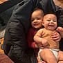 baby, infant, child, smile, diaper, red_sweater, beanbag, hand, indoor, wooden_floor, table, carpet, couch, person, face, happy, cute, sibling, holding, cozy