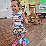 blue_floor_marker, child, clogs, colorful_clothing, curly_hair, dress, furniture, girl, happy, indoors, medical_office, pink_shoes, playful, portrait, rocking_chair, smiling, standing, toddler, waiting_room, wooden_floor