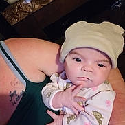 Rayna is registered to the contest to win money with this photo: baby, infant, hat, pajamas, monkey_pattern, hand, face, person, adult_arm, tattoo, tank_top, indoor, holding, expression, clothing, cute, newborn, skin, portrait, serious