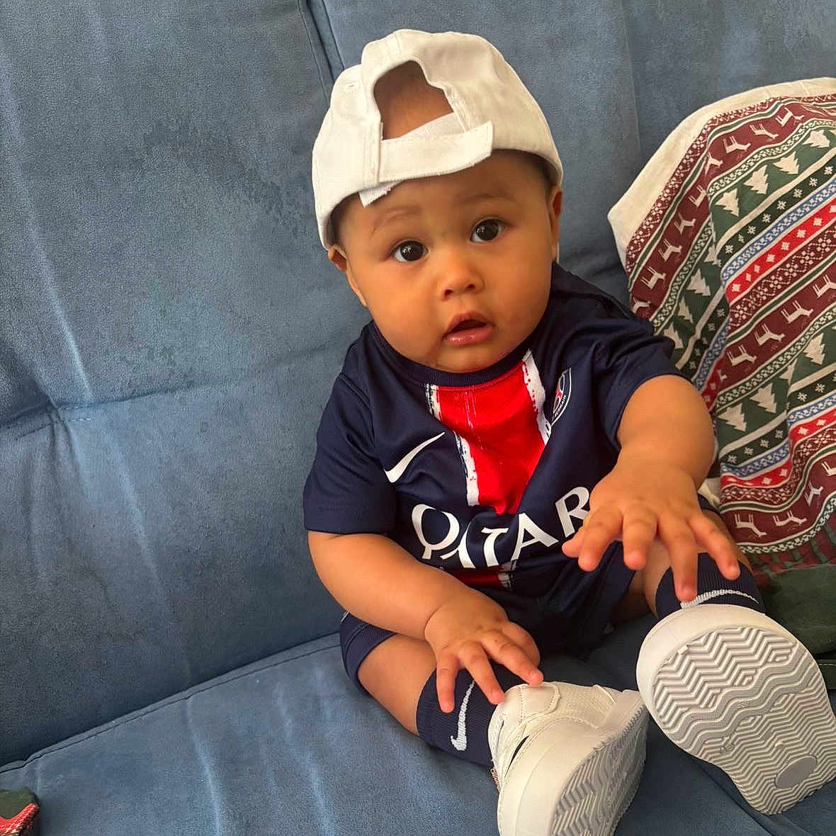 Neithan participe au concours pour gagner de l'argent avec cette photo : baby, blue_couch, cap, child, clothing, comfortable, couch, curious, footwear, hand, home, indoor, jersey, person, portrait, sitting, sneakers, sporty, wide_eyes, young
