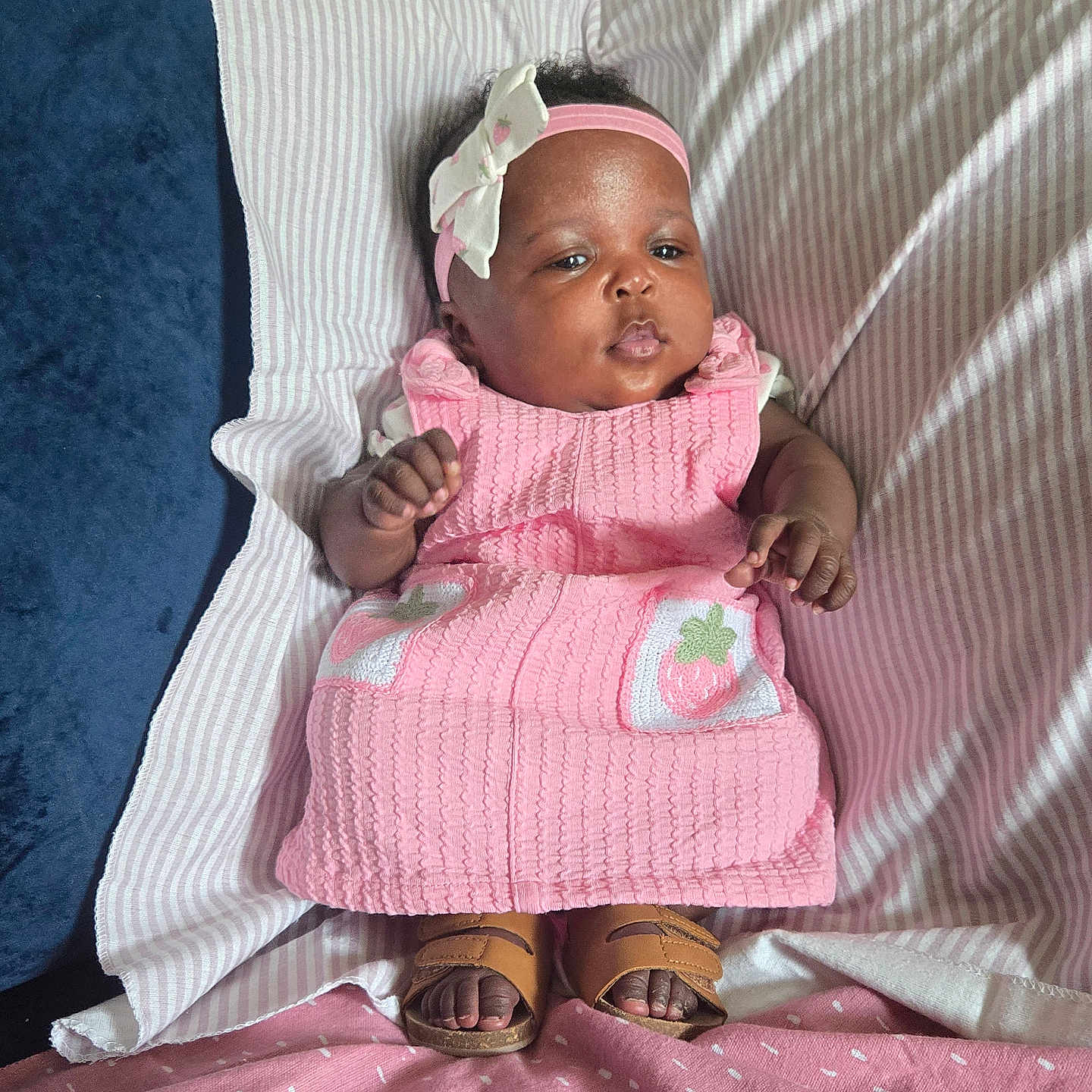 Wynter is registered to the contest to win money with this photo: baby, infant, child, portrait, lying_down, pink_dress, headband, bow, sandals, pillow, blanket, bed, indoor, smiling, fashion, cute, dark_skin, striped_fabric, strawberry, full_body