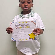 Taizir is registered to the contest to win money with this photo: achievement, award, beige_pants, boy, casual_clothing, certificate, child, colorful, education, holding, indoor, kindergarten, paper, person, portrait, proud, short_hair, smile, standing, white_shirt