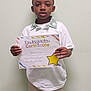 achievement, award, beige_pants, boy, casual_clothing, certificate, child, colorful, education, holding, indoor, kindergarten, paper, person, portrait, proud, short_hair, smile, standing, white_shirt
