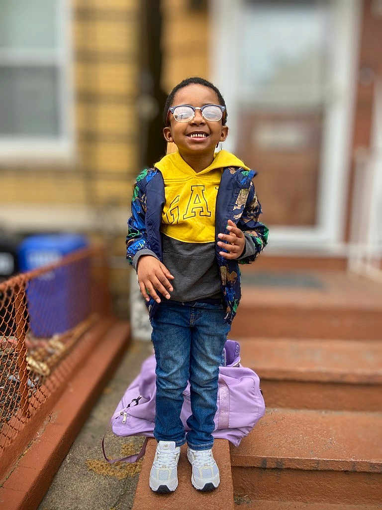 Taizir is registered to the contest to win money with this photo: child, boy, glasses, yellow_hoodie, jacket, jeans, sneakers, backpack, stairs, sidewalk, smile, outdoor, urban, fence, person, happy, casual_clothing, daylight, portrait, street