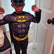 Taizir is registered to the contest to win money with this photo: child, batman_costume, mask, smile, indoor, door, cape, flexing, happy, muscles, floor, plastic_drawer, towel, foot, person, costume, superhero, toy, black_clothing, yellow_logo