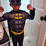 child, batman_costume, mask, smile, indoor, door, cape, flexing, happy, muscles, floor, plastic_drawer, towel, foot, person, costume, superhero, toy, black_clothing, yellow_logo