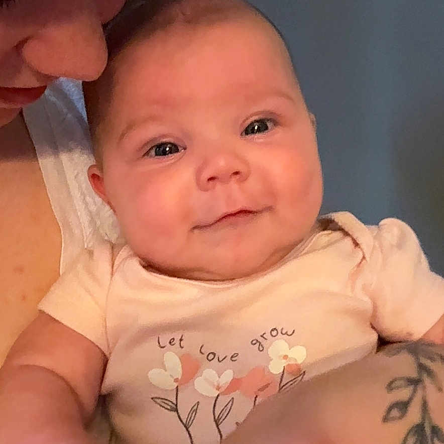 Ryker is registered to the contest to win money with this photo: adult, affection, arm, baby, closeup, clothing, face, flower, happy, holding, indoor, infant, love, onesie, person, portrait, skin, smile, tattoo, text