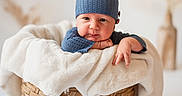 Ashton is registered to the contest to win money with this photo: baby, blue_hat, blue_sweater, basket, blanket, cute, face, greenery, infant, indoors, knitwear, neutral_background, person, plant, portrait, resting, soft_texture, white_blanket, woven_basket, young_child