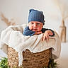 baby, blue_hat, blue_sweater, basket, blanket, cute, face, greenery, infant, indoors, knitwear, neutral_background, person, plant, portrait, resting, soft_texture, white_blanket, woven_basket, young_child