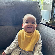 Helena is registered to the contest to win money with this photo: baby, bright, child, cozy, cute, furniture, gray_couch, happy, home_decor, indoor, infant, long_sleeve_clothes, natural_light, patterned_clothes, portrait, sitting, smile, window, yellow_bib, young_child