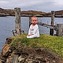 baby, calm, child, clothing, cloudy_sky, coast, daytime, grass, infant, landscape, nature, outdoor, quiet, rocks, scenic, serious_face, shore, sitting, water, wooden_posts