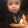Mathias joined the competition — help win amazing prizes! toddler, child, curly_hair, black_shirt, gold_embroidery, holding_papers, carpet, indoor, brown_eyes, smiling, standing, furniture, wooden, sneakers, cute, portrait, closeup, person, young_child, curious