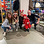 group, holiday, festive, child, toddler, red_coat, hat, reindeer_ears, santa_hat, smiling, glasses, store, clothing_rack, winter_jacket, shopping, indoor, people, floor, casual, joyful