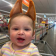 Elsie is registered to the contest to win money with this photo: toddler, child, shopping_cart, store, aisle, animal_ears, headband, smiling, clothing, striped_sweater, flower, indoor, person, cute, baby, face, happy, retail, shopping, cart