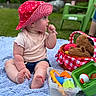 baby, blue_blanket, chewing, child, curious, cute, daylight, garden, grass, green_chair, outdoor, picnic, pink_shirt, plastic_toys, plush_toy, red_hat, sitting, soft_texture, summer, toy_basket