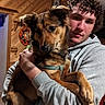 person, dog, indoor, hoodie, curly_hair, brown_dog, pet, holding, wooden_wall, casual, portrait, calm, young_adult, medium_dog, animal, companion, home, cozy, looking, face