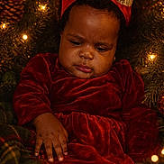 Brielle joined the competition — help win amazing prizes! baby, child, christmas_lights, closeup, cozy, cute, festive, gold_bow, greenery, headband, holiday, holiday_decor, indoor, pine_cone, plush, portrait, red_dress, serious_expression, toddler, warm_lighting