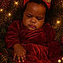 baby, child, christmas_lights, closeup, cozy, cute, festive, gold_bow, greenery, headband, holiday, holiday_decor, indoor, pine_cone, plush, portrait, red_dress, serious_expression, toddler, warm_lighting