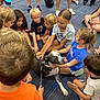 blue_shirt, carpet, casual_clothing, children, curious, diverse, dog, friendly, glasses, group, happy, indoor, interaction, leash, orange_shirt, petting, playful, shorts, sitting, young