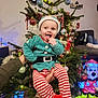 baby, child, christmas_tree, holiday, festive_clothing, elf_costume, striped_pants, red_booties, indoor, decorations, ornaments, lights, smiling, tongue_out, person, holding_hands, cheerful, seasonal, celebration, home