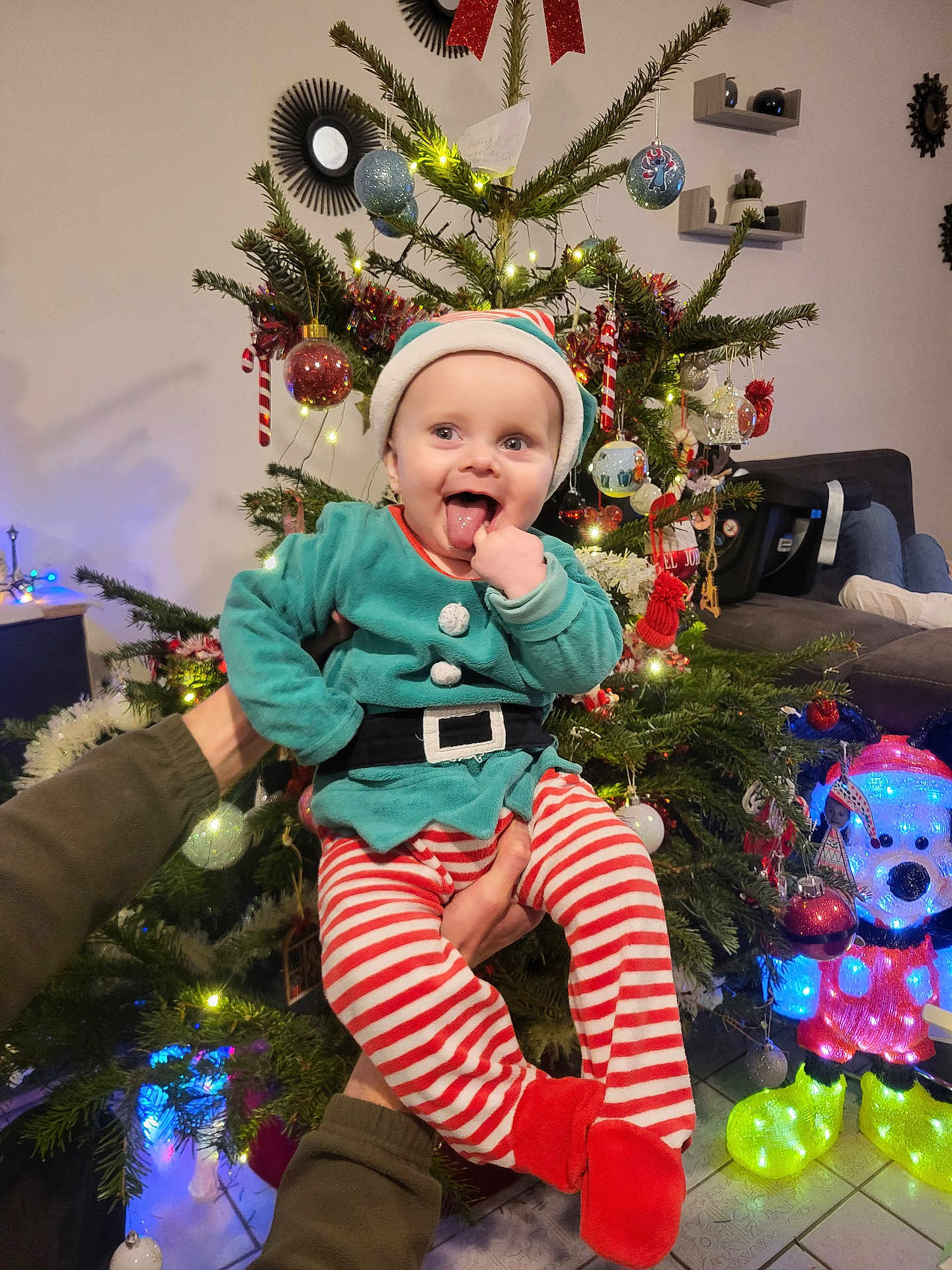 Néo participe au concours pour gagner de l'argent avec cette photo : baby, child, christmas_tree, holiday, festive_clothing, elf_costume, striped_pants, red_booties, indoor, decorations, ornaments, lights, smiling, tongue_out, person, holding_hands, cheerful, seasonal, celebration, home