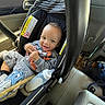 baby, child, infant, smiling, car_seat, car, vehicle, indoors, portrait, bottle, blanket, cartoon, clothing, gray_sweatshirt, sitting, harness, travel, road_trip, happy, automobile